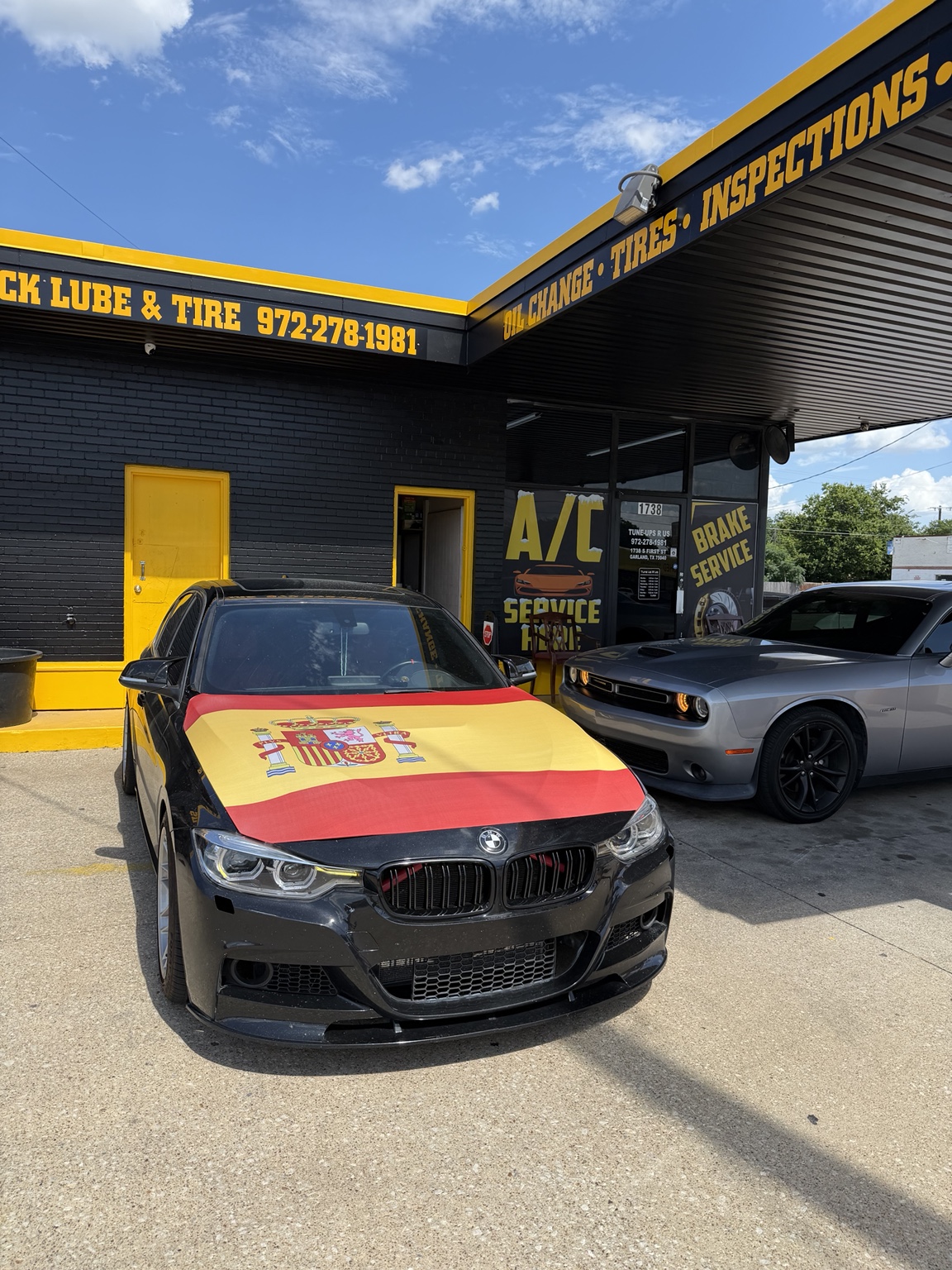 Black BMW 340i with a Spanish flag hood cover parked in front of the Tune Ups R Us shop
